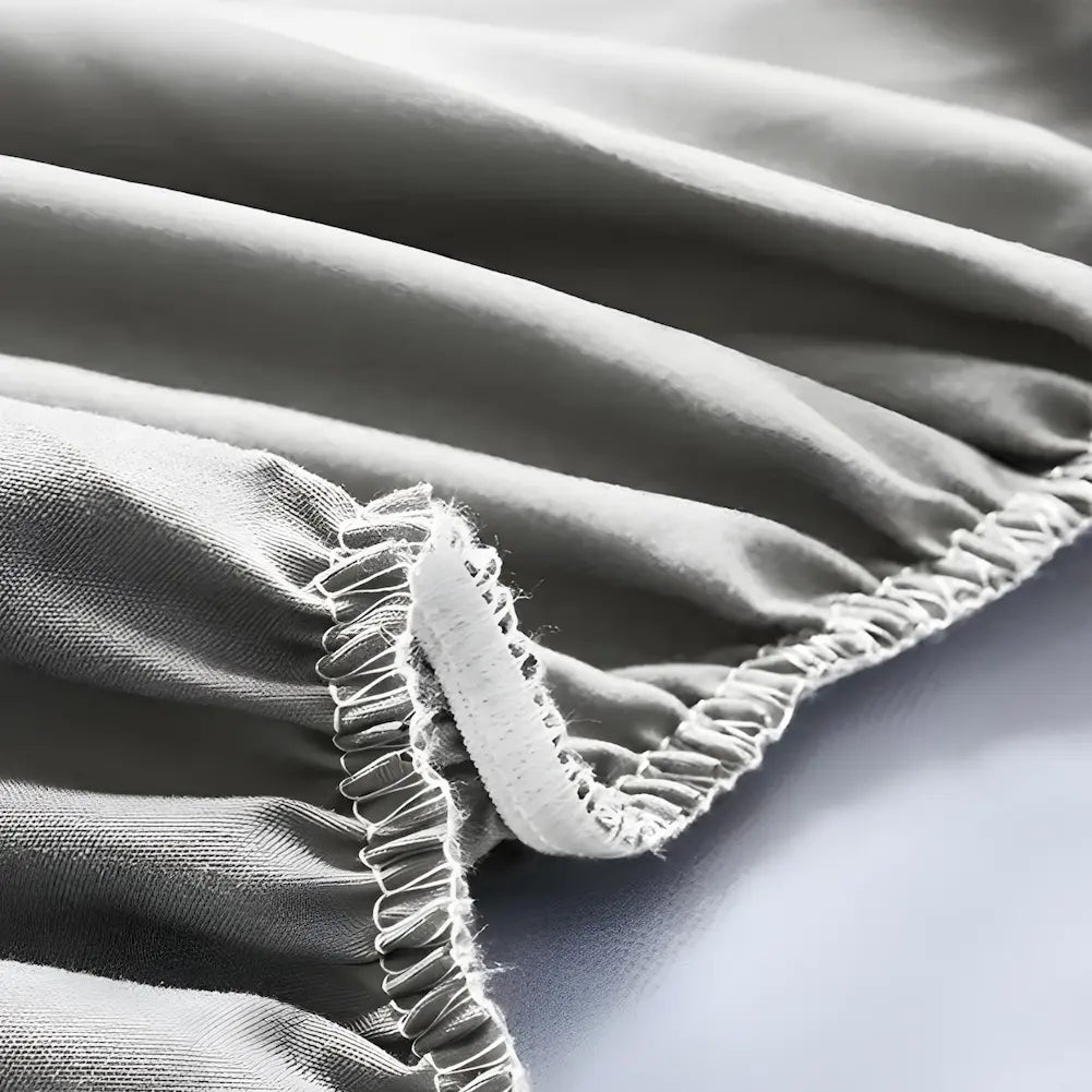Imagem em close de uma capa de colchão cinza, focada em sua borda elástica. A capa mostra costuras brancas reforçadas, detalhando o elástico que permite ajuste firme ao colchão. O tecido parece ser macio e ligeiramente acolchoado, com um aspecto suave e uniforme.